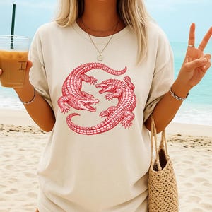 May include: Cream-colored t-shirt featuring a red illustration of two alligators facing each other. The alligators are detailed with scales and claws. The shirt is a relaxed fit, suitable for casual wear. The design is centered on the front of the shirt.