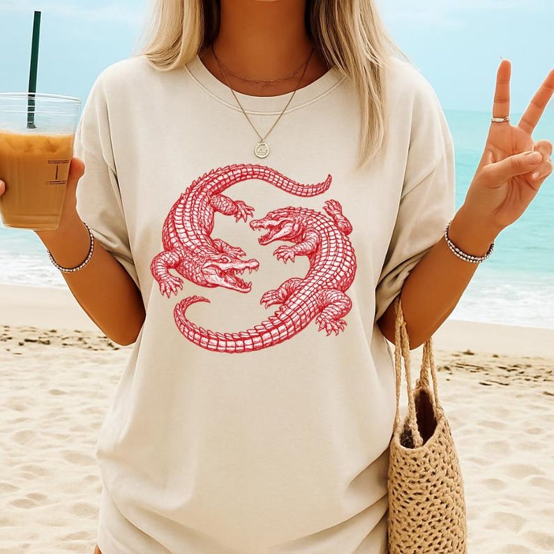 May include: Cream-colored t-shirt featuring a red illustration of two alligators facing each other. The alligators are detailed with scales and claws. The shirt is a relaxed fit, suitable for casual wear. The design is centered on the front of the shirt.
