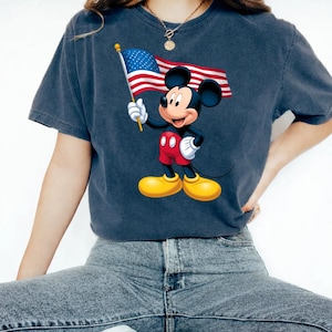 Puede incluir: Camiseta azul oscuro con un gr&aacute;fico de Mickey Mouse sosteniendo una bandera estadounidense. Mickey lleva pantalones cortos rojos, zapatos amarillos y guantes blancos. La bandera tiene rayas rojas y blancas y un campo azul con estrellas blancas.