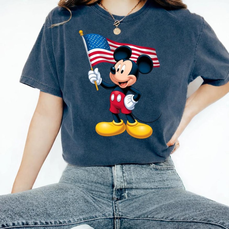 Puede incluir: Camiseta azul oscuro con un gr&aacute;fico de Mickey Mouse sosteniendo una bandera estadounidense. Mickey lleva pantalones cortos rojos, zapatos amarillos y guantes blancos. La bandera tiene rayas rojas y blancas y un campo azul con estrellas blancas.