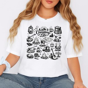 May include: White t-shirt featuring a black and white illustration of camping-related items. The design includes a campfire, tent, lantern, backpack, guitar, and the words "Camping Life".