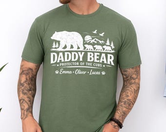 May include: An olive green short-sleeve t-shirt featuring a white graphic of a large bear, three smaller bears, mountains, and trees. The text reads DADDY BEAR PROTECTOR OF THE CUBS with the names Emma, Oliver, and Lucas printed below with paw prints.