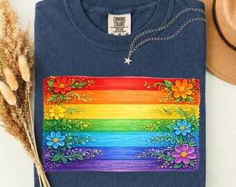 May include: A navy blue t-shirt featuring a rectangular graphic of a rainbow-colored horizontal striped background decorated with painted flowers and leaves in the corners. The shirt has a white inner neck label with the text Comfort Colors.