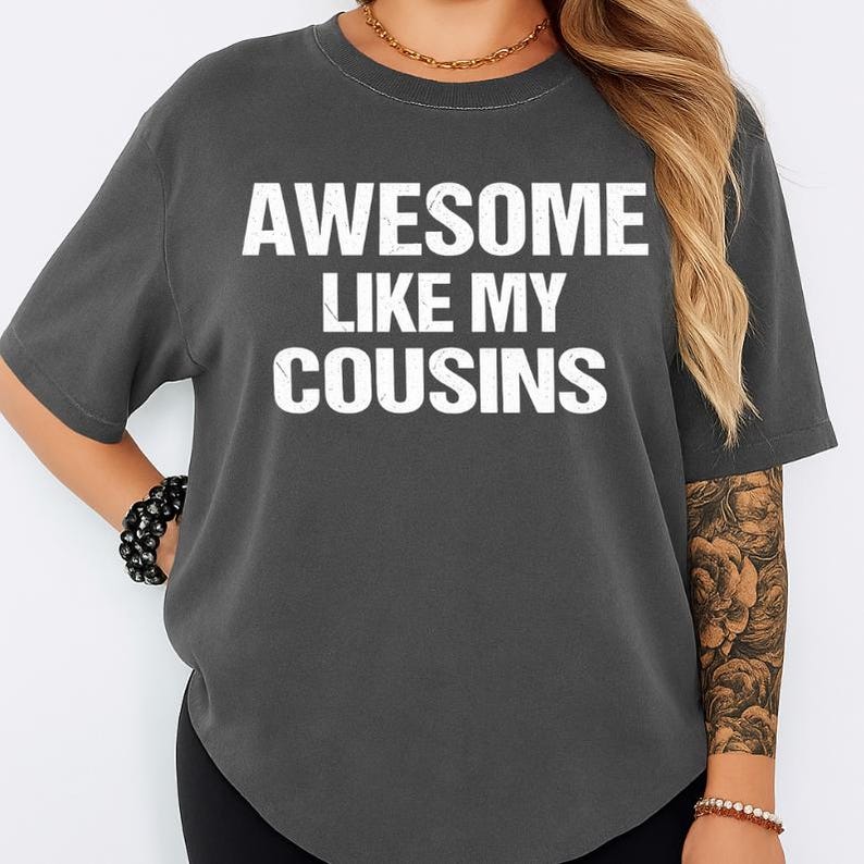 May include: Dark gray t-shirt with the text "AWESOME LIKE MY COUSINS" in white block letters. The shirt has short sleeves and a crew neck. The shirt is made of a soft material.