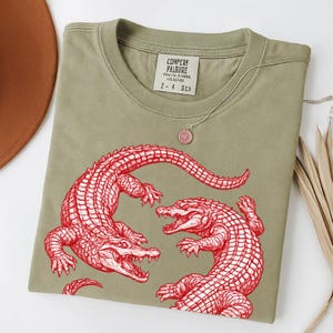 May include: An olive green t-shirt featuring a red illustration of two alligators facing each other. The shirt has a crew neck and a tag with text at the neckline. A silver necklace with a pink pendant is draped on the shirt.