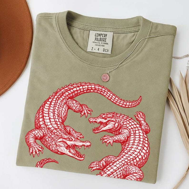 May include: An olive green t-shirt featuring a red illustration of two alligators facing each other. The shirt has a crew neck and a tag with text at the neckline. A silver necklace with a pink pendant is draped on the shirt.