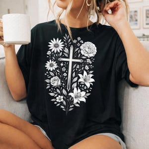 Op de afbeelding: Zwart t-shirt met een bloemenkruisontwerp. De afbeelding bevat een kruis omringd door witte bloemen, waaronder madeliefjes, lelies en rozen. Het ontwerp is gecentreerd op de voorkant van het shirt.