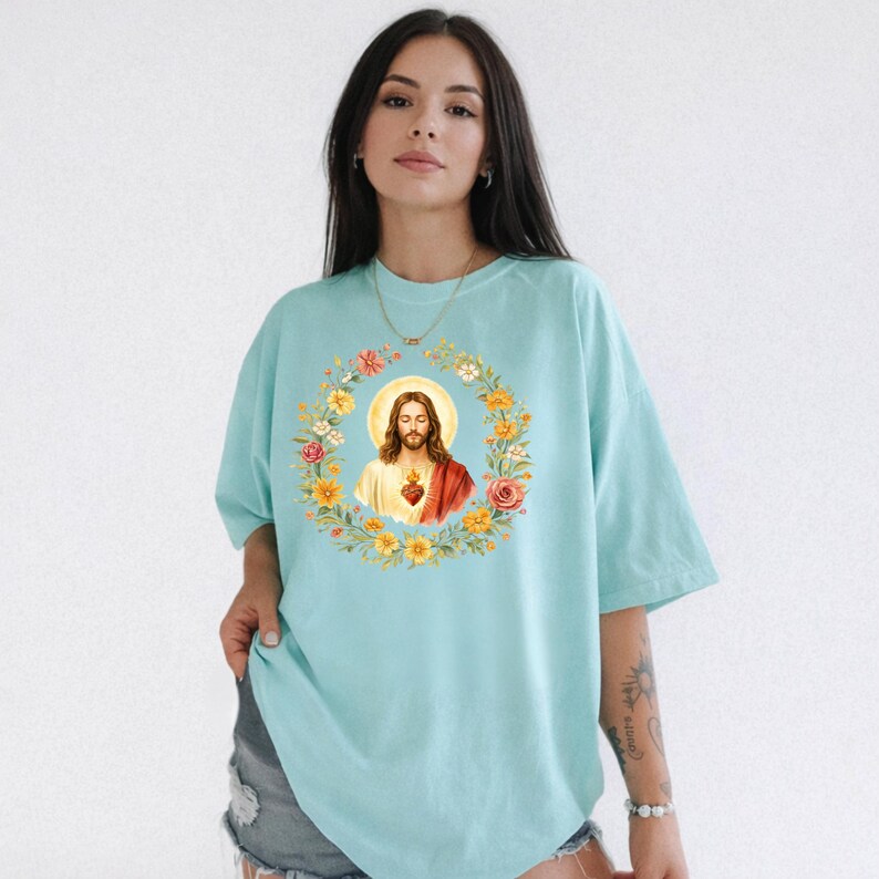 K&ouml;nnte beinhalten: Ein hellblaues T-Shirt mit einer zentralen Grafik von Jesus Christus mit Heiligenschein, umgeben von einem kreisf&ouml;rmigen Kranz aus bunten Blumen, darunter gelbe, orangefarbene und rosa Bl&uuml;ten. Das Motiv ist auf der Vorderseite gedruckt.
