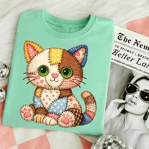 May include: A light green crew neck t-shirt featuring a patchwork cat design. The cat has a white face, brown and yellow ears, and a brown body with colorful patches. The cat has large green eyes and pink paws.