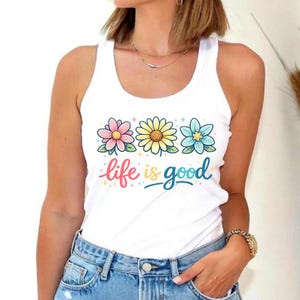 May include: White tank top with a floral design featuring three flowers in pink, yellow, and blue. The words "life is good" are written in colorful script below the flowers. The top is made of a soft material.