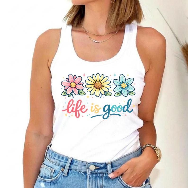 May include: White tank top with a floral design featuring three flowers in pink, yellow, and blue. The words "life is good" are written in colorful script below the flowers. The top is made of a soft material.