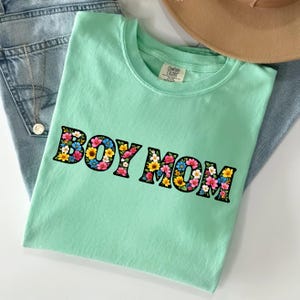 May include: A mint green t-shirt with the text "BOY MOM" in a floral design. The letters are outlined in black and filled with colorful flowers. The shirt is folded on top of blue jeans.