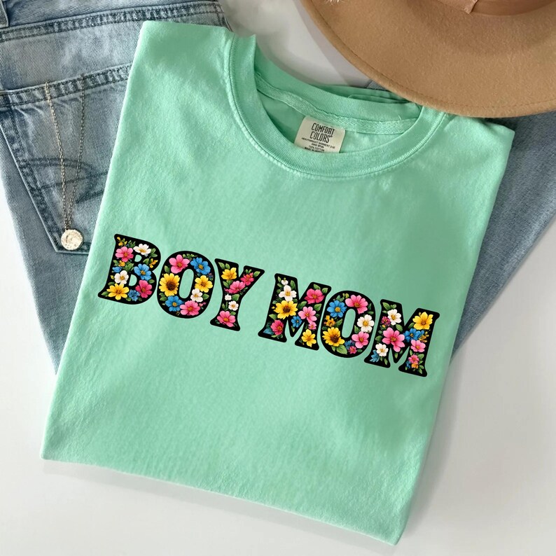 May include: A mint green t-shirt with the text "BOY MOM" in a floral design. The letters are outlined in black and filled with colorful flowers. The shirt is folded on top of blue jeans.