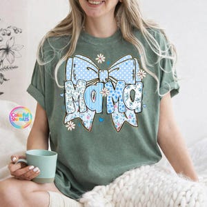 May include: Green t-shirt with a large graphic that says "Mama" in blue floral and polka dot letters, surrounded by a blue bow and small white flowers. The shirt has rolled-up sleeves and the text "Colorful ShirtsUS" is visible.