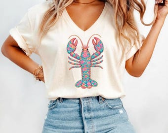 May include: Cream-colored V-neck t-shirt featuring a colorful lobster graphic. The lobster design incorporates pink, blue, and yellow floral patterns. The shirt is paired with blue denim jeans.