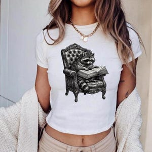 Könnte beinhalten: Ein weißes Kurzarm-T-Shirt mit einer schwarz-weißen grafischen Illustration eines Waschbären, der in einem gepolsterten Sessel sitzt und ein offenes Buch liest. Das Motiv ist mittig auf der Brust platziert und zeigt detaillierte Linienführung.