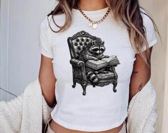 May include: A white short-sleeved t-shirt featuring a black and white graphic illustration of a raccoon sitting in a tufted armchair while reading an open book. The design is centered on the chest area of the garment, showcasing detailed line work.