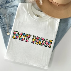 May include: A white t-shirt with the text "BOY MOM" in a floral design. The letters are outlined in black and filled with colorful flowers. The shirt is laying on a pair of blue jeans.