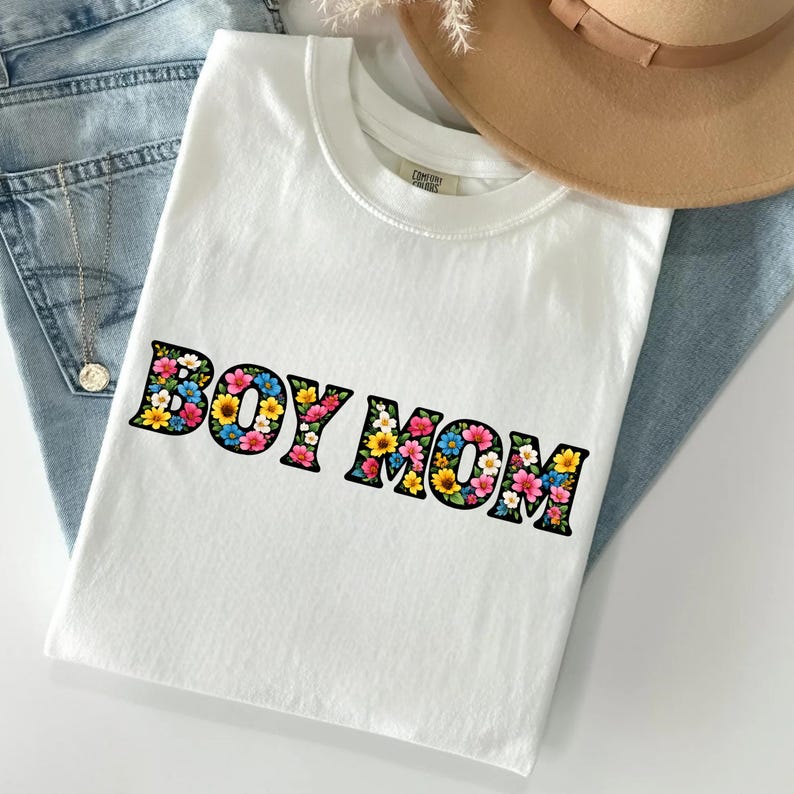May include: A white t-shirt with the text "BOY MOM" in a floral design. The letters are outlined in black and filled with colorful flowers. The shirt is laying on a pair of blue jeans.