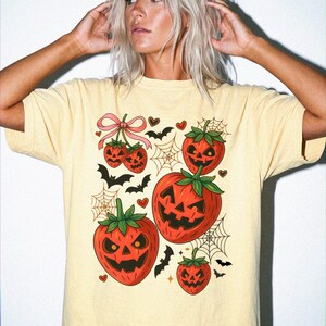 Könnte beinhalten: Ein hellgelbes Kurzarm-T-Shirt mit einem Grafikdruck von mehreren Erdbeeren, die wie Kürbisse geschnitzt sind. Das Design enthält kleine schwarze Fledermäuse, Spinnennetze, winzige rote Herzen und eine rosa Schleife um zwei der Erdbeeren.