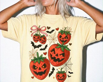 May include: A pale yellow short-sleeve t-shirt featuring a graphic print of multiple strawberries carved like jack-o'-lanterns. The design includes small black bats, spider webs, tiny red hearts, and a pink ribbon tied around two strawberries.