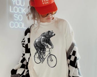 May include: Cream-colored t-shirt featuring a black and white illustration of a bear riding a bicycle. The image also shows a red trucker hat with the text "MAD LOVE FOR YOU". The person is wearing a black and white checkered cardigan.