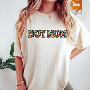 May include: Cream-colored t-shirt with the text "BOY MOM" in a floral design. The letters are filled with colorful flowers, including yellow, blue, and pink. The shirt has short sleeves and a relaxed fit.