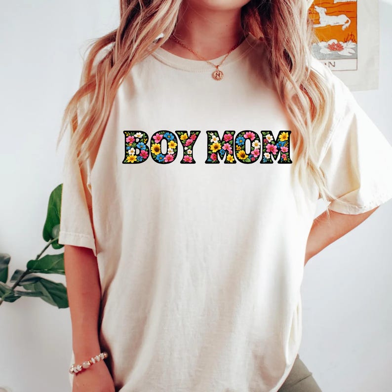 May include: Cream-colored t-shirt with the text "BOY MOM" in a floral design. The letters are filled with colorful flowers, including yellow, blue, and pink. The shirt has short sleeves and a relaxed fit.