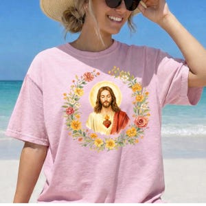 K&ouml;nnte beinhalten: Ein hellrosa T-Shirt mit einem floralen Kranzdesign und einem zentralen Bild von Jesus. Das Jesusbild hat lange braune Haare, ein wei&szlig;es und rotes Gewand und einen leuchtenden Heiligenschein. Der Kranz enth&auml;lt gelbe und rosa Blumen.