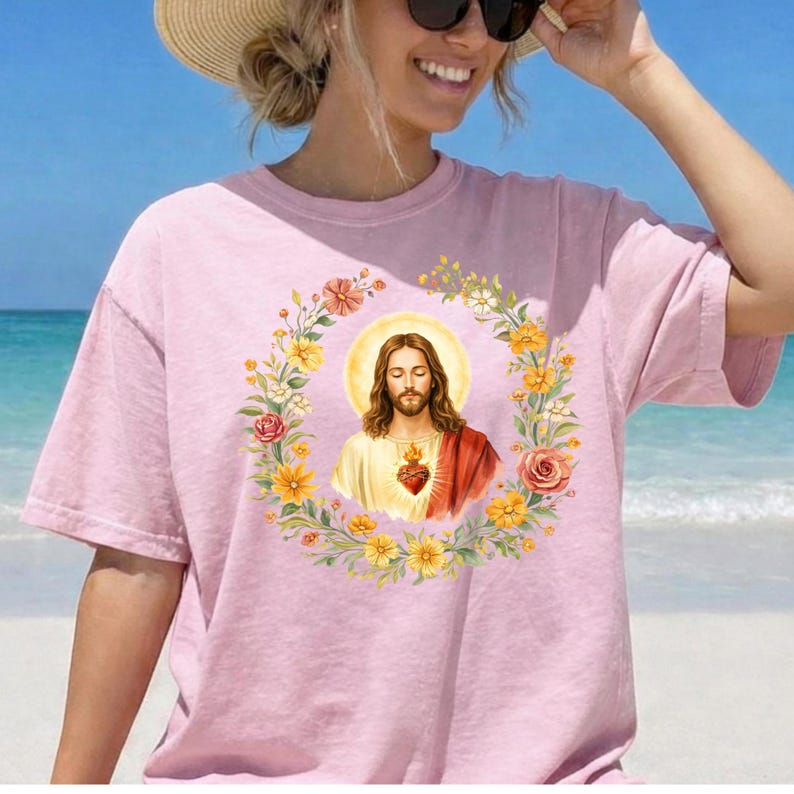 K&ouml;nnte beinhalten: Ein hellrosa T-Shirt mit einem floralen Kranzdesign und einem zentralen Bild von Jesus. Das Jesusbild hat lange braune Haare, ein wei&szlig;es und rotes Gewand und einen leuchtenden Heiligenschein. Der Kranz enth&auml;lt gelbe und rosa Blumen.