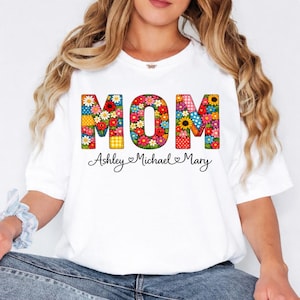 May include: White t-shirt featuring the word "MOM" in colorful floral patchwork letters. Below the word "MOM" are the names "Ashley Michael Mary" in a cursive font.