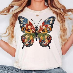 May include: Cream-colored t-shirt featuring a large, colorful butterfly design. The butterfly has intricate patterns in shades of blue, red, orange, and green. The shirt is a casual style, suitable for everyday wear.