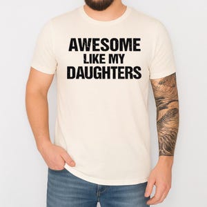 May include: Cream-colored t-shirt with the text "AWESOME LIKE MY DAUGHTERS" in bold black letters. The shirt is short-sleeved and worn by a person wearing blue jeans. The person has a beard and a tattoo on their arm.