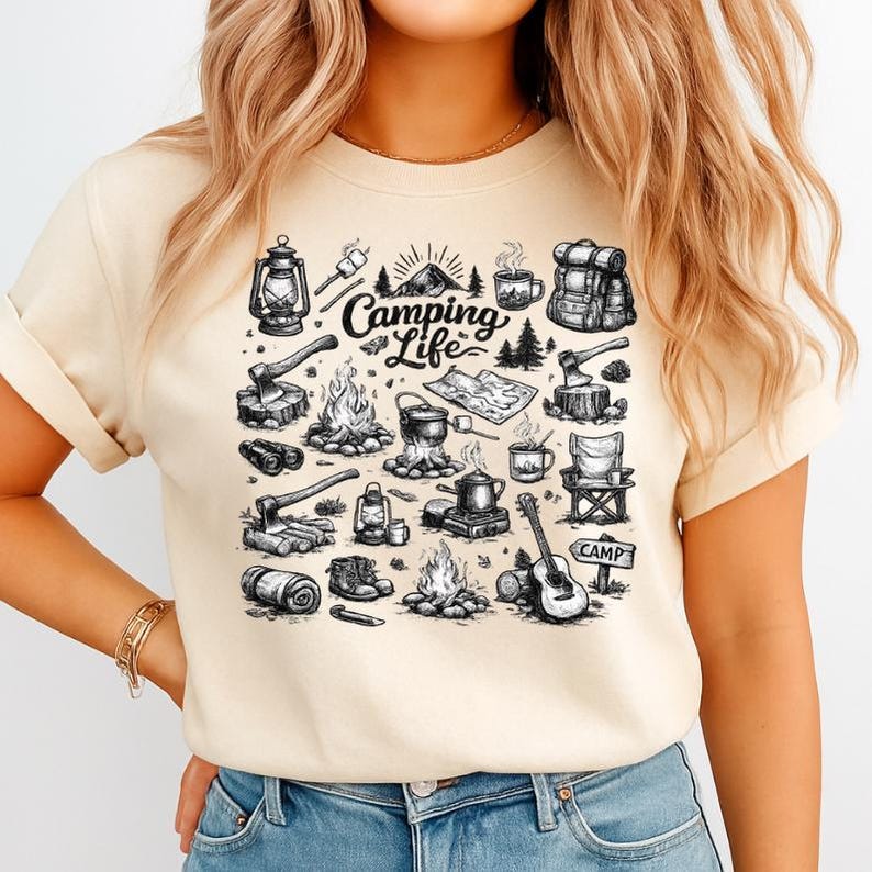 May include: Beige t-shirt with a black and white camping-themed graphic. The design includes illustrations of a lantern, campfire, backpack, axe, and the words "Camping Life". A guitar and a sign that says "Camp" are also featured.