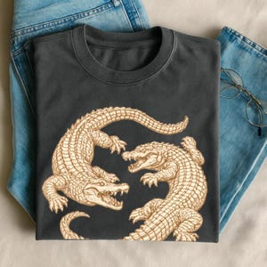 May include: Dark gray t-shirt featuring a detailed illustration of two alligators facing each other. The alligators are tan with textured scales, set against a dark gray background. The shirt is folded on top of blue jeans.