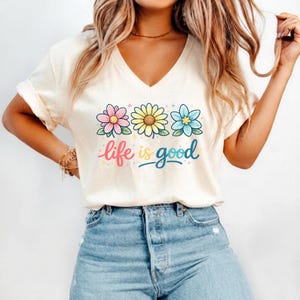 May include: Cream-colored V-neck t-shirt featuring three colorful flowers and the phrase "life is good" in a whimsical font. The flowers are pink, yellow, and blue, with green leaves. The shirt is paired with blue jeans.