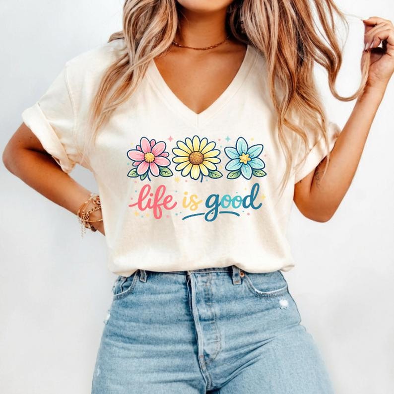 May include: Cream-colored V-neck t-shirt featuring three colorful flowers and the phrase "life is good" in a whimsical font. The flowers are pink, yellow, and blue, with green leaves. The shirt is paired with blue jeans.