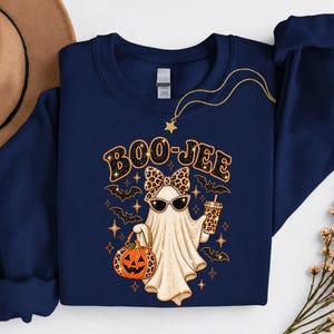 Pu&ograve; includere: Una felpa blu navy a maniche lunghe con la grafica di un fantasma che indossa un fiocco e occhiali da sole leopardati, mentre tiene in mano un secchiello a forma di zucca e una bevanda. Il testo BOO-JEE &egrave; stampato sopra, circondato da pipistrelli.