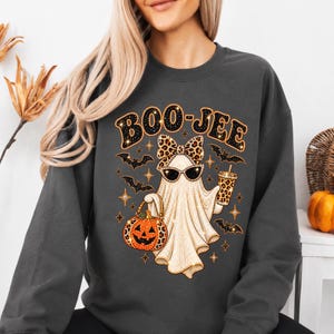 Pu&ograve; includere: Una felpa girocollo grigio scuro con la grafica di un fantasma che indossa occhiali da sole e un fiocco leopardato. Il fantasma tiene in mano un secchiello a forma di zucca e un bicchiere leopardati, circondato da pipistrelli e la scritta BOO-JEE.