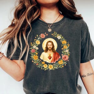 K&ouml;nnte beinhalten: Dunkelgraues T-Shirt mit einem Blumenkranz, der ein Bild von Jesus mit Heiligenschein umgibt. Das Bild zeigt Jesus mit einem roten Herzen und einem wei&szlig;en und roten Gewand. Der Kranz enth&auml;lt gelbe, rosa und wei&szlig;e Blumen.