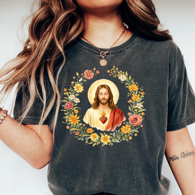 K&ouml;nnte beinhalten: Dunkelgraues T-Shirt mit einem Blumenkranz, der ein Bild von Jesus mit Heiligenschein umgibt. Das Bild zeigt Jesus mit einem roten Herzen und einem wei&szlig;en und roten Gewand. Der Kranz enth&auml;lt gelbe, rosa und wei&szlig;e Blumen.