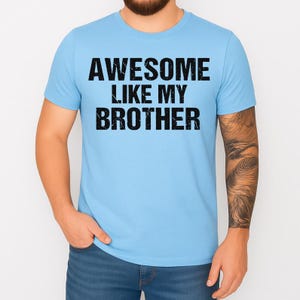 May include: Light blue t-shirt with the text "AWESOME LIKE MY BROTHER" in bold, black letters. The shirt is short-sleeved and appears to be made of a soft material. The person is wearing blue jeans.