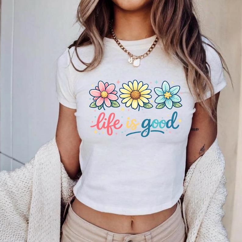 May include: White t-shirt with a colorful floral design and the phrase "life is good." The graphic features three flowers in pink, yellow, and blue, with green stems. The text is in a playful, handwritten style.