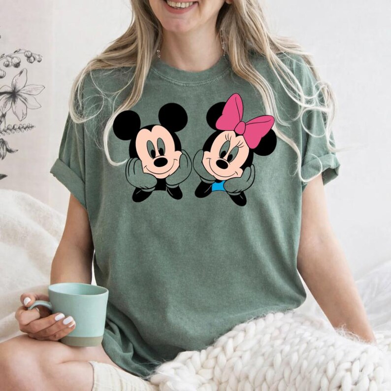 May include: Sage green t-shirt featuring a graphic of Mickey and Minnie Mouse. Mickey has a black bow tie and Minnie has a pink bow. The cartoon characters are smiling and resting their chins on their hands.