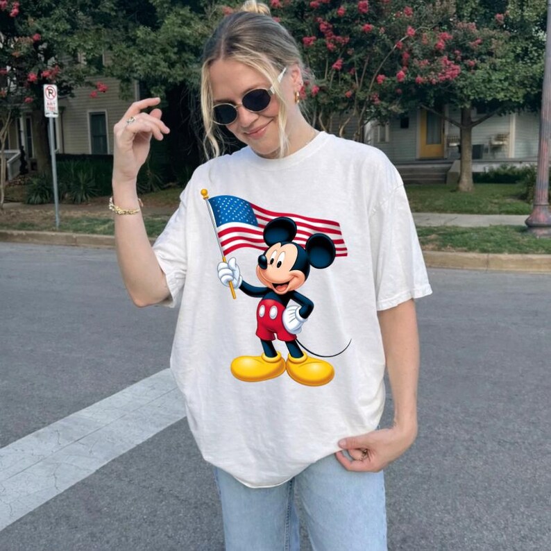 Puede incluir: Camiseta blanca con un dibujo de Mickey Mouse sosteniendo una bandera estadounidense. Mickey lleva pantalones cortos rojos, zapatos amarillos y guantes blancos. La bandera tiene rayas rojas y blancas y un campo azul con estrellas blancas.