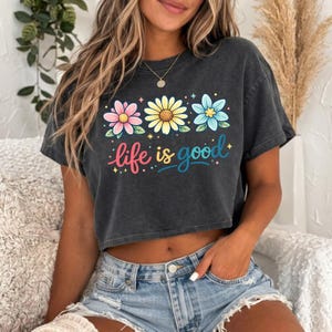May include: A dark gray crop top with a floral design and the phrase "life is good" in colorful script. The top features three flowers in pink, yellow, and blue. The shirt is paired with denim shorts.