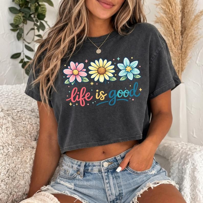 May include: A dark gray crop top with a floral design and the phrase "life is good" in colorful script. The top features three flowers in pink, yellow, and blue. The shirt is paired with denim shorts.