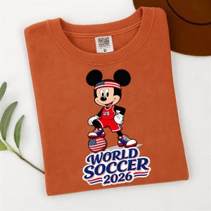 May include: An orange t-shirt featuring a graphic of Mickey Mouse in a soccer uniform with a US flag headband. The design includes a soccer ball and the text "WORLD SOCCER 2026".