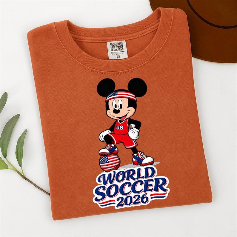 May include: An orange t-shirt featuring a graphic of Mickey Mouse in a soccer uniform with a US flag headband. The design includes a soccer ball and the text "WORLD SOCCER 2026".