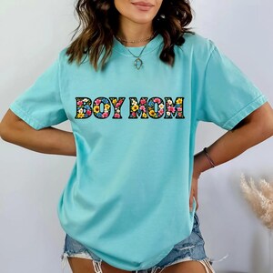 May include: A light blue t-shirt with the text "BOY MOM" in a floral design. The shirt has short sleeves and a relaxed fit. The text is in a bold, block letter style with a colorful flower pattern.